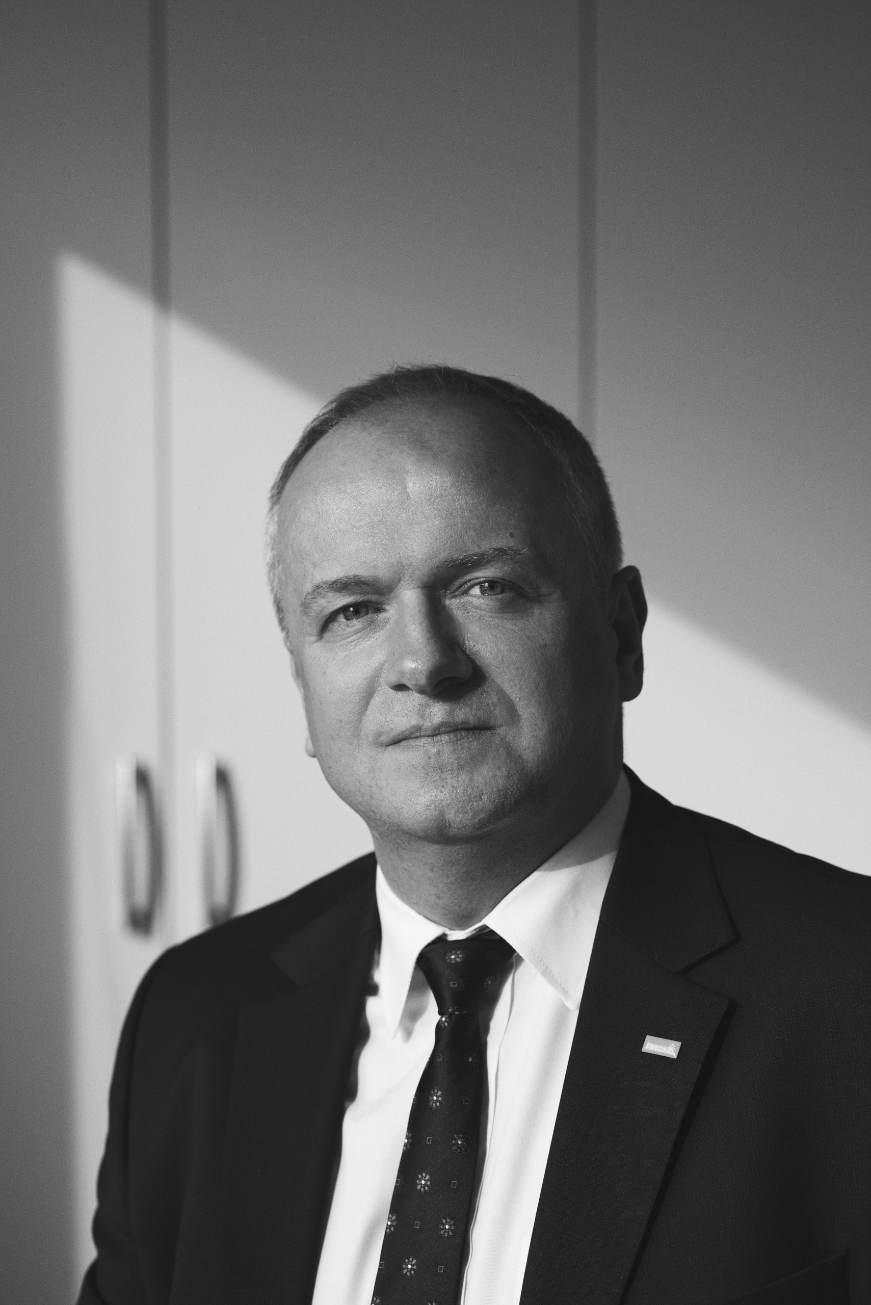 Czarno-biały, profesjonalny portret wizerunkowy mężczyzny w garniturze i krawacie, patrzącego prosto w obiektyw. Ma krótkie włosy i stoi na tle ściany z wyraźnym cieniem, co dodaje zdjęciu eleganckiej, korporacyjnej głębi.