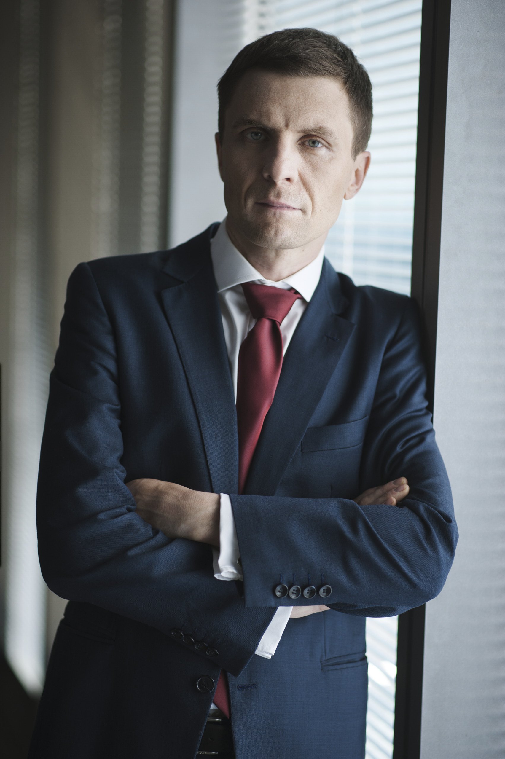 Mężczyzna w garniturze z czerwonym krawatem pozuje do profesjonalnego portretu biznesowego, z założonymi rękami, stojąc przy oknie z żaluzjami; miękkie światło z okna modeluje twarz, a tło pozostaje stonowane i neutralne.